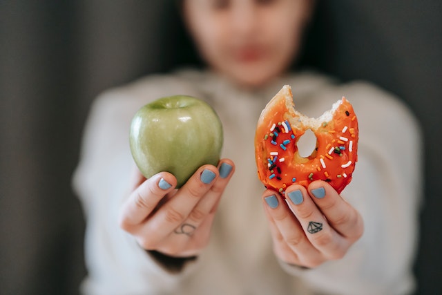 A woman holding a green apple and a bitten piece of donut with icing and candy sprinkles.