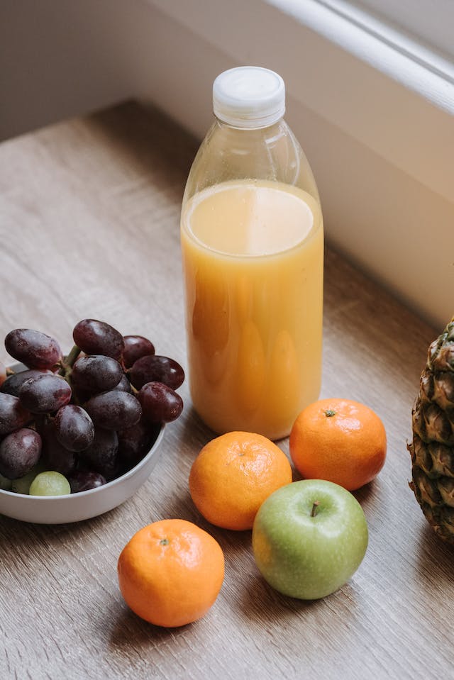 A bowl of grapes, three oranges, one green apple, and a bottle of fruit juice placed on a wooden table.