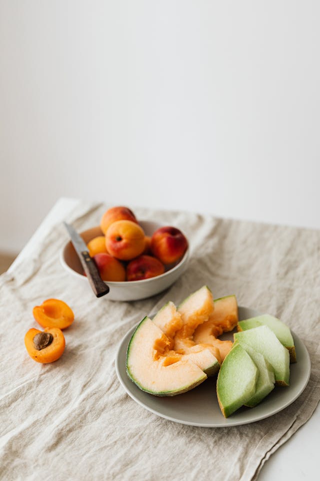 A serving of sliced fresh melons and a bowl of apricots placed on a wooden table.