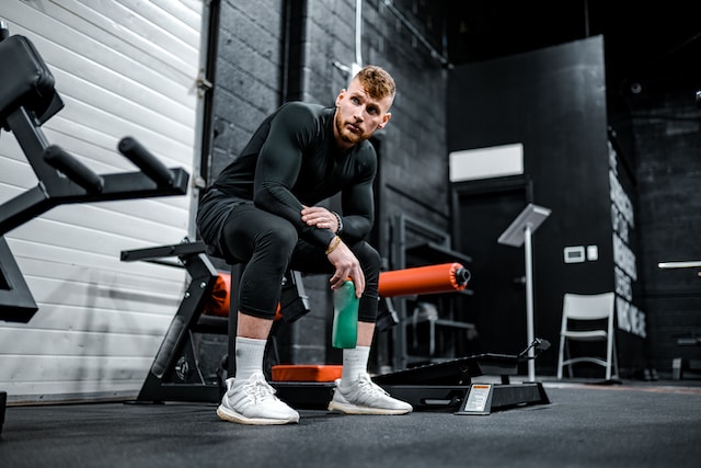 A man wearing black sports attire and holding a green bottle sitting on exercise equipment