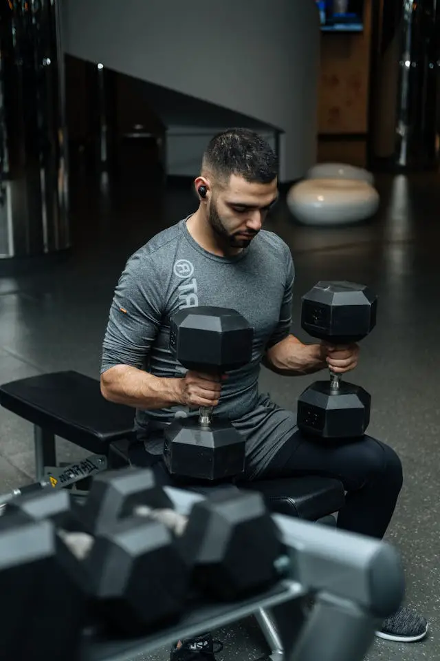 A man sitting on a gym bench while holding two heavy dumbbells on his lap.