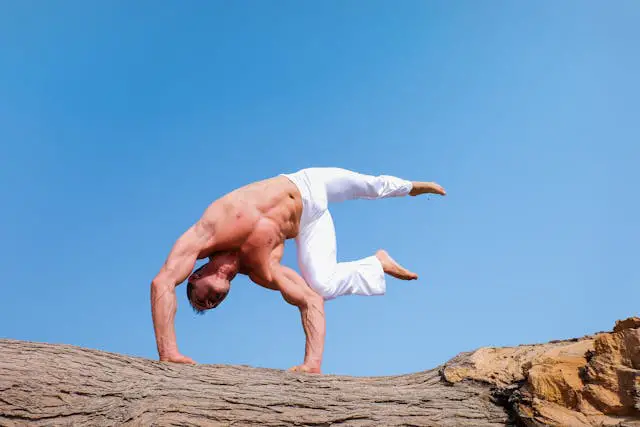 A shirtless muscular man in white pants tumbling on a big trunk under the clear blue skies.