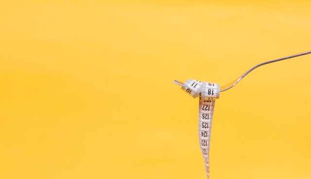 A close-up of a fork with a measuring tape twirled around it, set against a bright yellow background