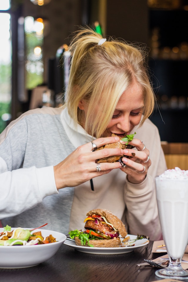 A woman happily bites into a burger, surrounded by plates of food and a milkshake