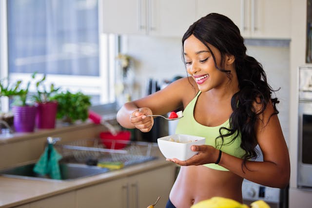 A smiling woman holding a bowl and about to eat spoonful of fruits in the kitchen.