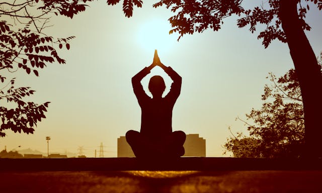 A silhoutte of a person meditating under the tree while facing the sun.