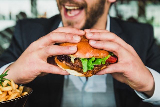 A man in a suit eagerly holds and prepares to bite into a burger, with a side of fries on the table.